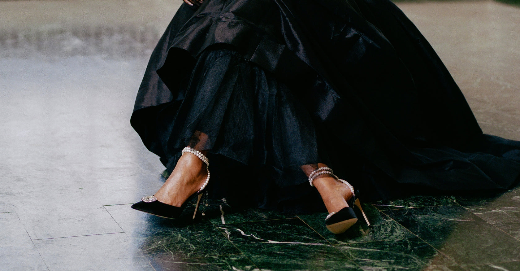 Zena Ziora luxury footwear capsule collection: The Dame pearl stiletto heel with crystal rectangular diamond embellishment in black satin. Model wearing shoes with legs crossed. Black formal ballgown in view.