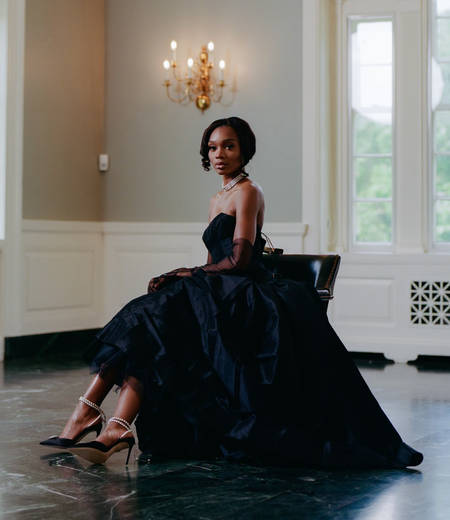 Model in black ball gown, gloves and necklace sitting down facing camera wearing Zena Ziora luxury footwear capsule collection: The Dame pearl stiletto heel with crystal rectangular diamond embellishment in black satin. 