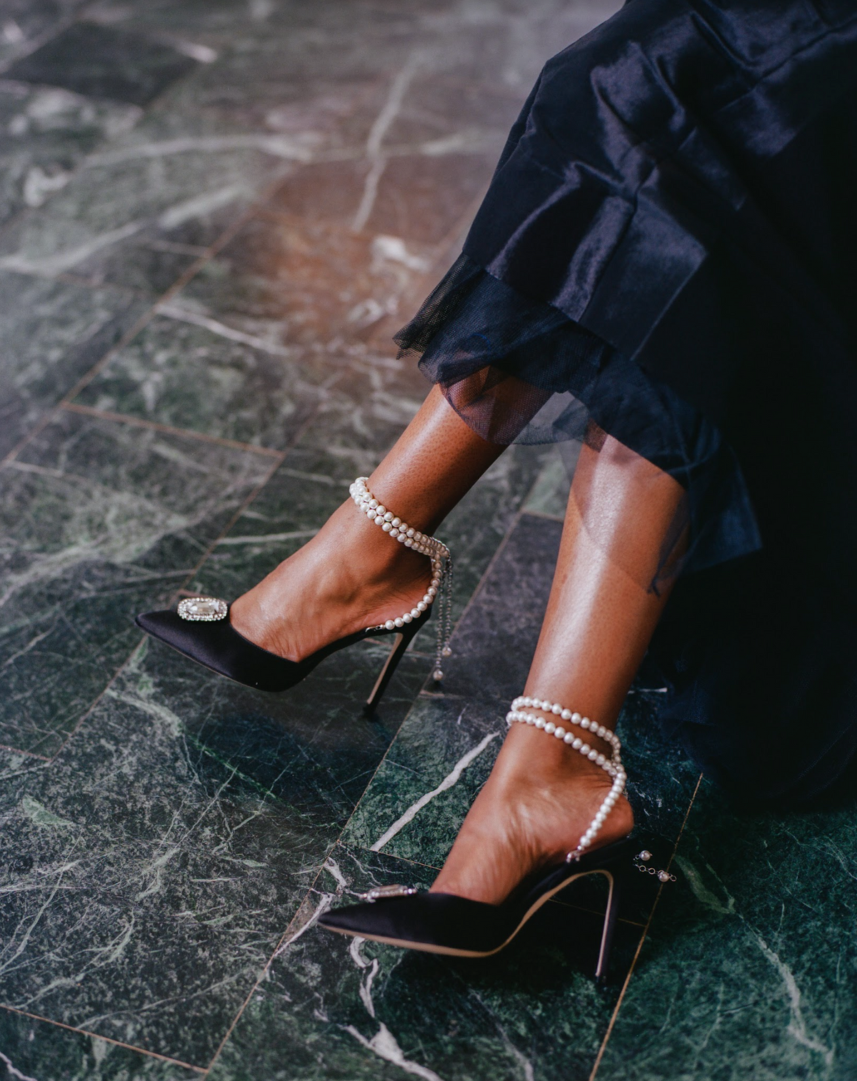 Zena Ziora luxury footwear capsule collection: The Dame pearl stiletto heel with crystal rectangular diamond embellishment in black satin. Model wearing shoes with legs crossed. Black formal ballgown in view.