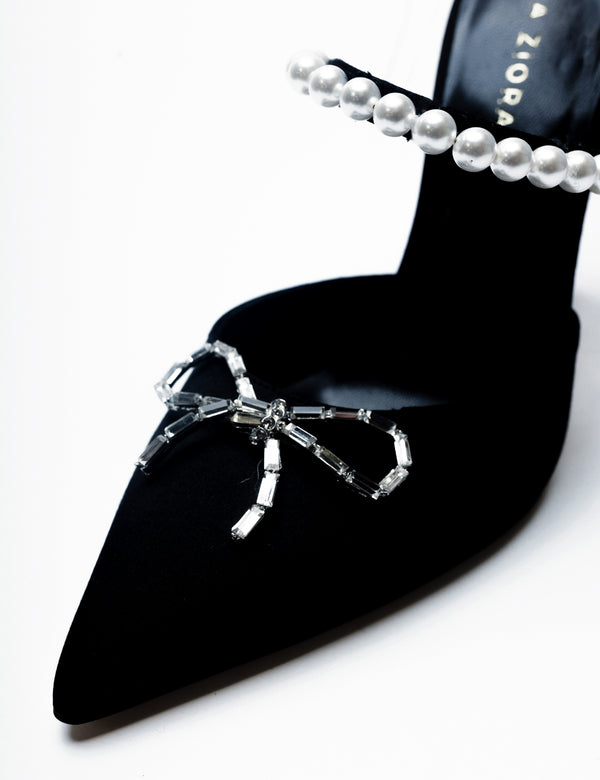 Zena Ziora luxury footwear capsule collection: Philippa pearl stiletto mule with crystal bow embellishment in black satin. Zoomed in detail shot displaying crystal bow and pearl strap on diagonal angle. 