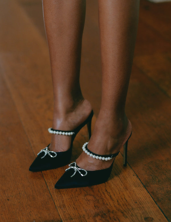Zena Ziora luxury footwear capsule collection: Philippa pearl stiletto mule with crystal bow embellishment in black satin. Model standing up wearing both shoes; legs shown. 