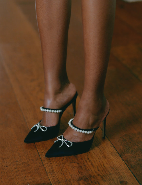 Zena Ziora luxury footwear capsule collection: Philippa pearl stiletto mule with crystal bow embellishment in black satin. Model standing up wearing both shoes; legs shown. 