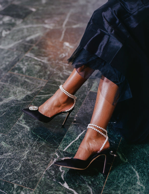 Zena Ziora luxury footwear capsule collection: The Dame pearl stiletto heel with crystal rectangular diamond embellishment in black satin. Model wearing shoes with legs crossed. Black ball gown in view.